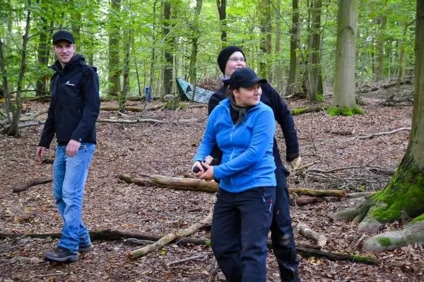 Drei Personen bewegen sich durch einen Wald mit Laub und Ästen am Boden