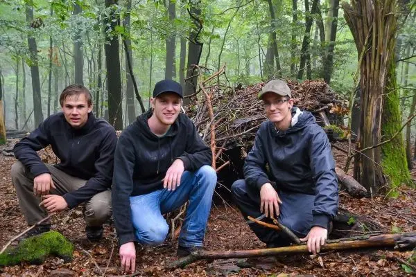 Drei Personen hocken vor einer selbstgebauten Laubhütte im Wald