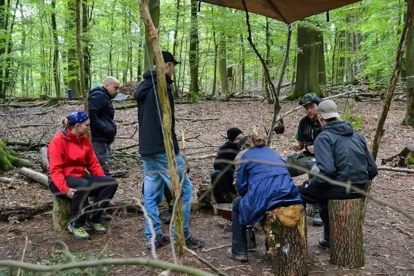 Gruppe sitzt auf Baumstümpfen unter einem Tarp im Wald und diskutiert