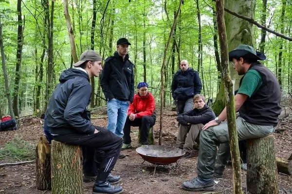 Gruppe sitzt um ein Feuer in einem Wald, Holzstämme als Sitzgelegenheiten