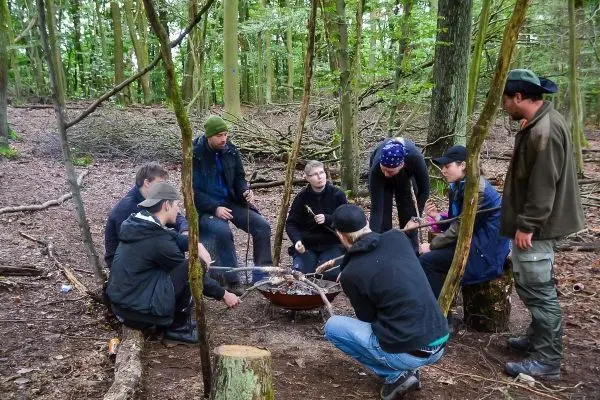 Gruppe sitzt um ein Feuer in einem Wald, Holzstücke und Töpfe sind sichtbar