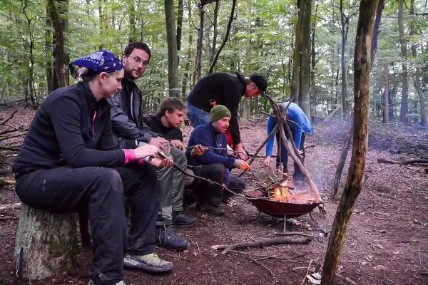 Gruppe sitzt um ein Feuer und röstet Essen auf Stöcken beim Survival Training