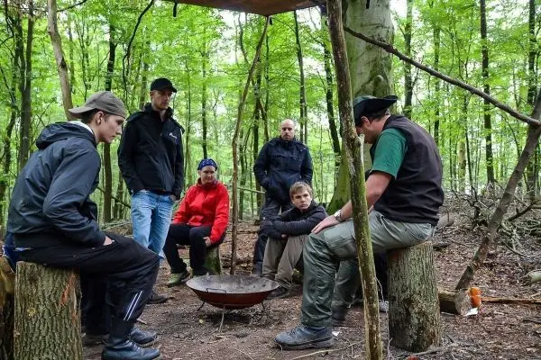 Gruppe sitzt um eine Feuerstelle im Wald und hört einem Mann zu beim Überlebenstraining