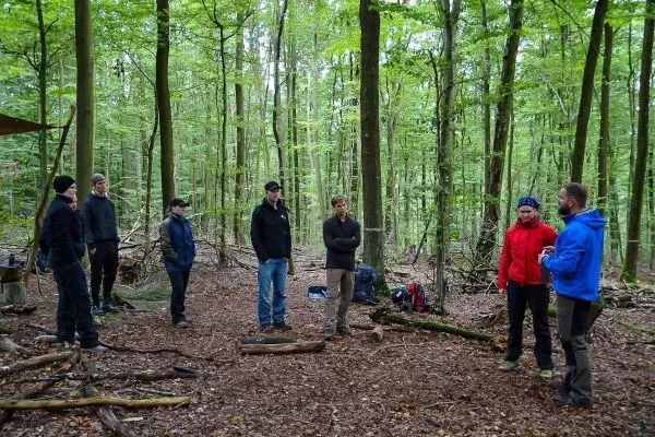 Gruppe steht im Wald um einen Holzstamm, diskutiert über Überlebenstechniken