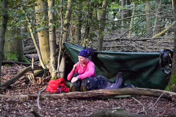 Person sitzt vor einer Tarp-Notunterkunft im Wald und hält einen Stock