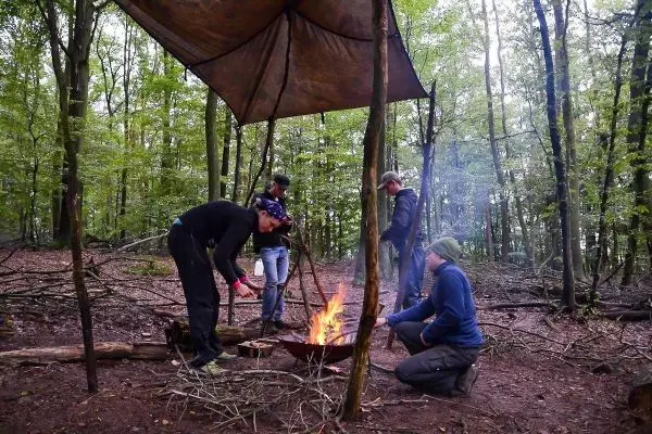 Personen stehen um ein Feuer unter einem Zelt im Wald