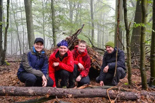 Vier Personen posieren vor einer selbstgebauten Tarp-Notunterkunft im Wald