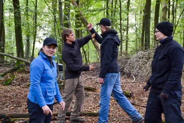 Vier Personen stehen im Wald und interagieren miteinander, während sie einen Stock halten
