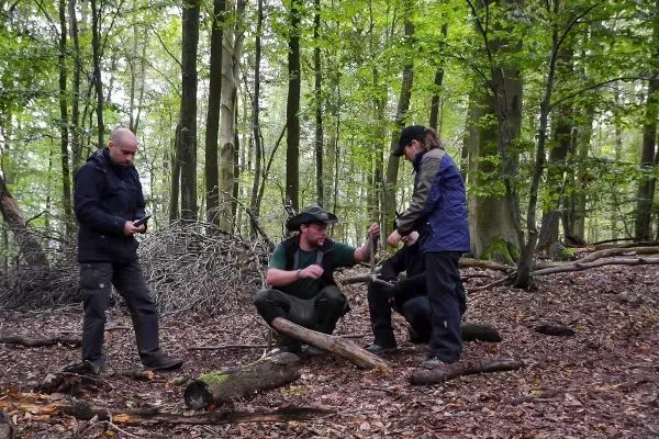 Vier Personen stehen und sitzen im Wald, während sie Holzstücke bearbeiten