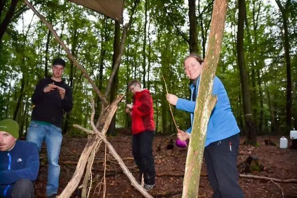 Zwei Personen bauen eine Holzstruktur im Wald, eine hält einen Stock