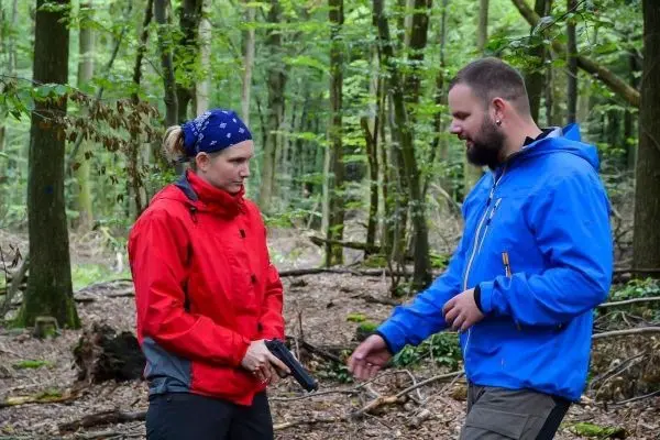 Zwei Personen im Wald, eine hält eine Pistole, der andere spricht mit ihr