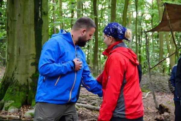 Zwei Personen in Outdoor-Bekleidung stehen im Wald und interagieren miteinander