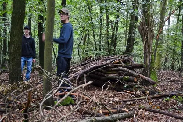 Zwei Personen stehen neben einem selbstgebauten Unterstand aus Ästen im Wald