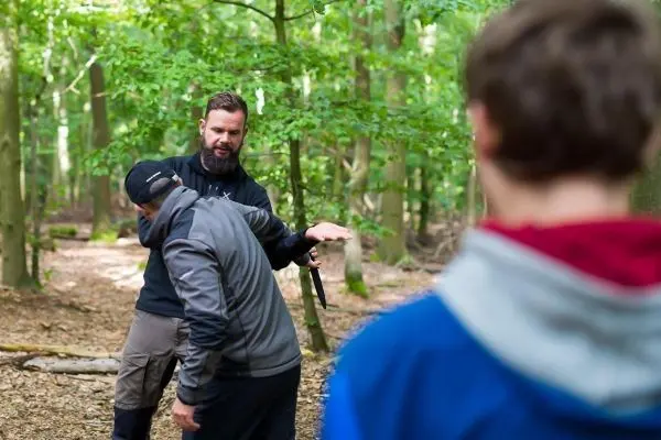 Ein Trainer demonstriert eine Technik im Wald, während zwei Teilnehmer aufmerksam zuschauen