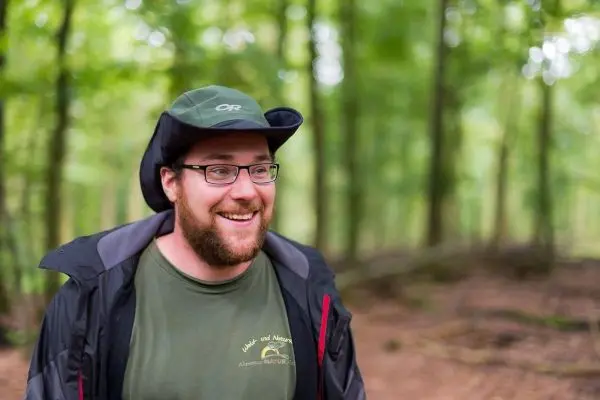 Lächelnder Mann mit Brille und Hut steht im Wald