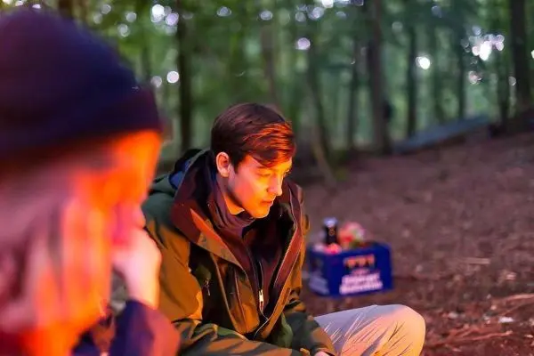 Lagerfeuer erhellt den Wald, zwei Personen sitzen am Boden und schauen nachdenklich