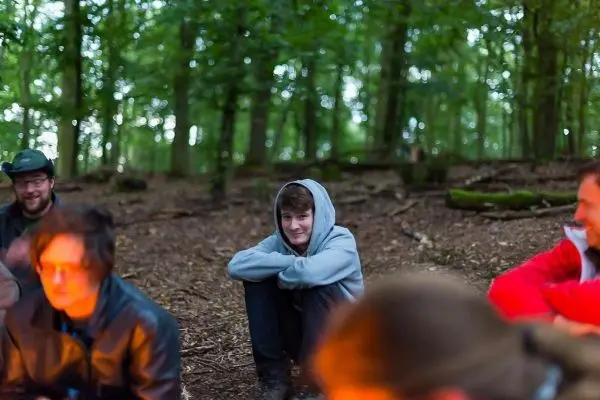 Lagerfeuer in einem Wald, mehrere Personen sitzen im Kreis und unterhalten sich