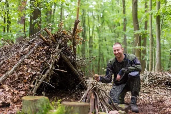 Mann kniet neben einer selbstgebauten Laubhütte im Wald
