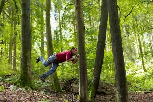 Mann überquert selbstgebaute Seilbrücke zwischen zwei Bäumen im Wald