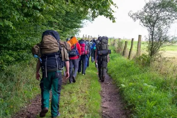 Wandernde Gruppe mit Rucksäcken auf einem schmalen Pfad entlang einer Wiese