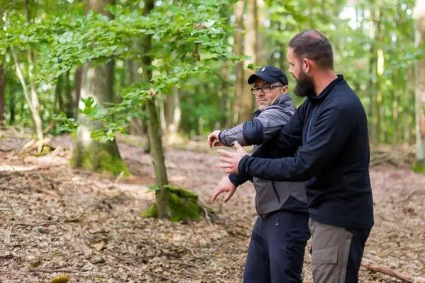 Zwei Männer demonstrieren eine Technik im Wald, umgeben von Bäumen und Laub