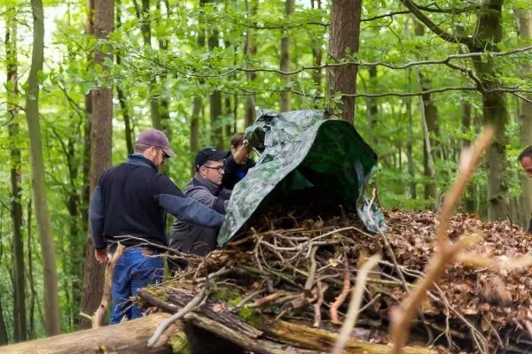 Zwei Personen befestigen eine Tarp-Notunterkunft auf einem Laubhaufen im Wald