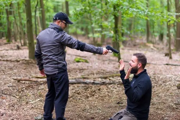 Zwei Personen im Wald, eine kniet nieder und hebt die Hände, während die andere eine Pistole hält