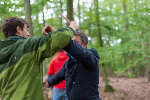 Zwei Personen im Wald üben eine Nahkampftechnik mit einem Stock
