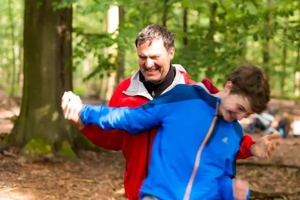 Zwei Personen interagieren spielerisch im Wald, umgeben von Bäumen und Laub