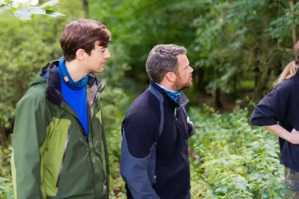 Zwei Personen stehen im Wald und schauen in Richtung einer dritten Person