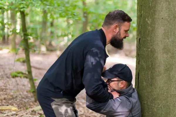 Zwei Personen stehen in einem Wald, eine Person hält die andere am Hals