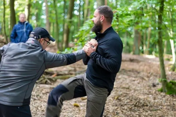 Zwei Personen üben eine Selbstverteidigungstechnik im Wald auf einem mit Laub bedeckten Boden