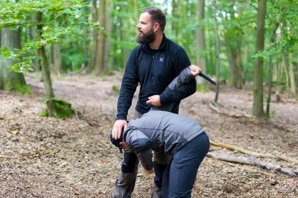 Zwei Personen üben eine Technik im Wald, einer hält ein Messer in der Hand