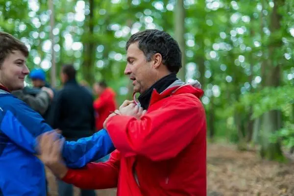 Zwei Personen üben eine Technik im Wald, einer trägt eine rote Jacke