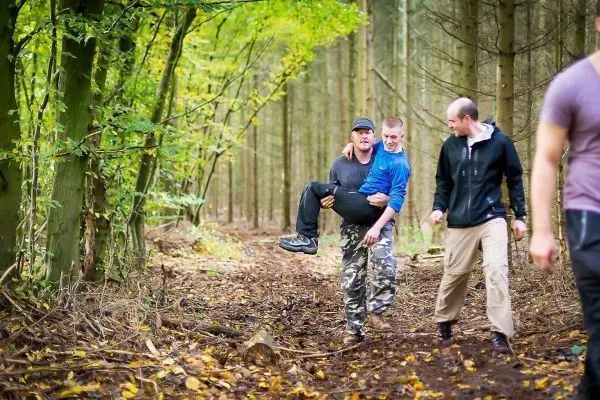 Drei Personen tragen einen verletzten Mann auf einem Waldweg