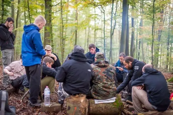Gruppe sitzt im Wald um einen Baumstamm und diskutiert beim Survival Training