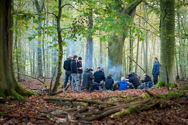 Gruppe sitzt um ein kleines Feuer im Wald, Rauch steigt auf