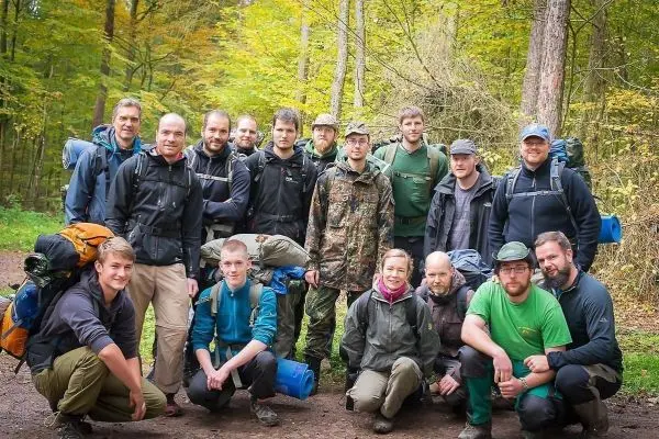 Gruppierung von Personen mit Rucksäcken im Wald, umgeben von Bäumen und herbstlichem Laub