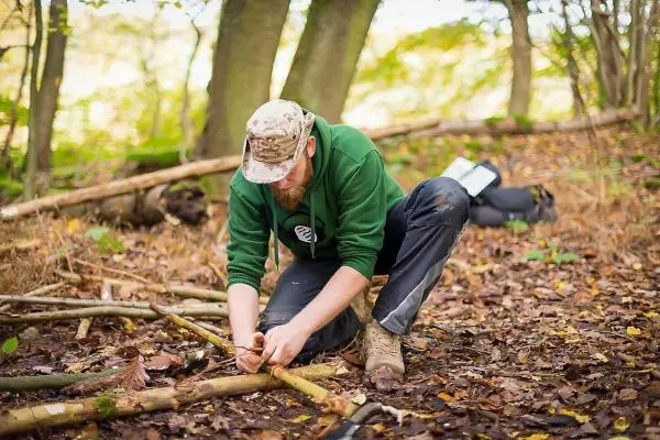 Holzstück wird mit einem Messer bearbeitet, während Laub und Äste im Hintergrund liegen