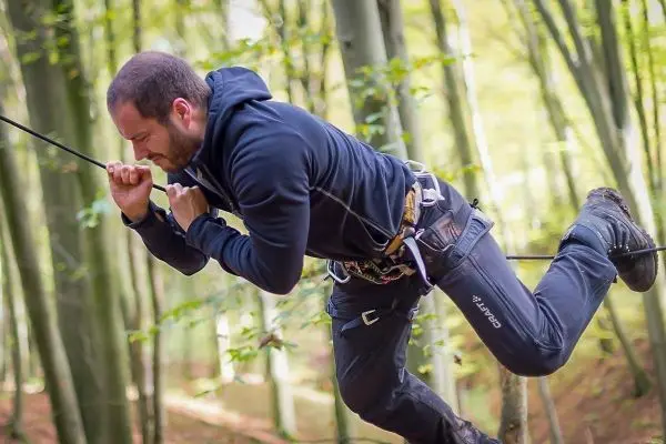 Mann überquert selbstgebaute Seilbrücke im Wald mit Sicherheitsgurt und Seil