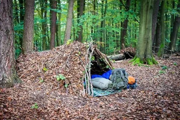 Tarp-Notunterkunft aus Ästen und Laub im Wald mit Rucksack davor