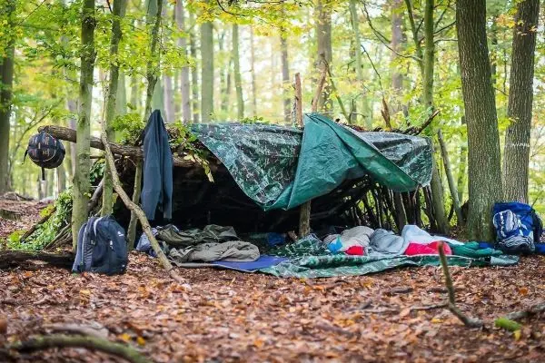 Tarp-Notunterkunft aus Ästen und Planen im Wald, Schlafsäcke und Rucksäcke liegen daneben