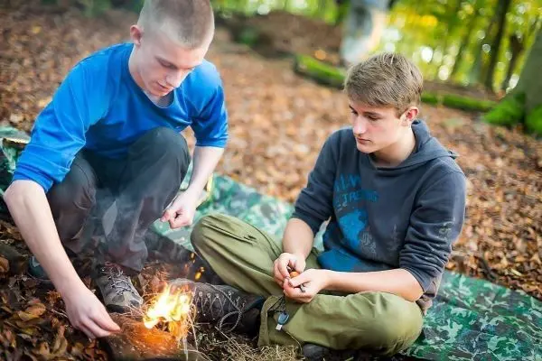 Zunder glimmt neben zwei Jugendlichen auf dem Boden im Wald