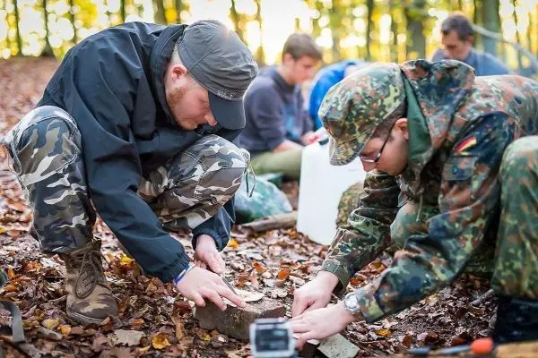 Zwei Personen bearbeiten einen Holzblock im Laub, während weitere Teilnehmer im Hintergrund arbeiten