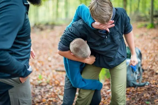 Zwei Personen in Outdoor-Bekleidung interagieren im Wald, eine Person umarmt die andere