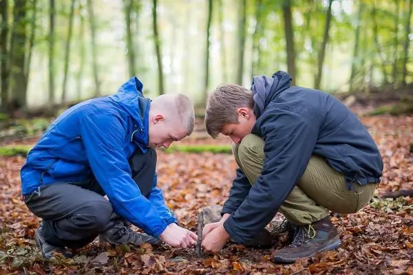 Zwei Personen knien im Laub und bearbeiten einen kleinen Holzstock