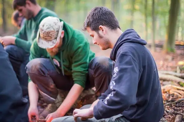 Zwei Personen sitzen im Wald und bearbeiten Holzstücke mit Messern