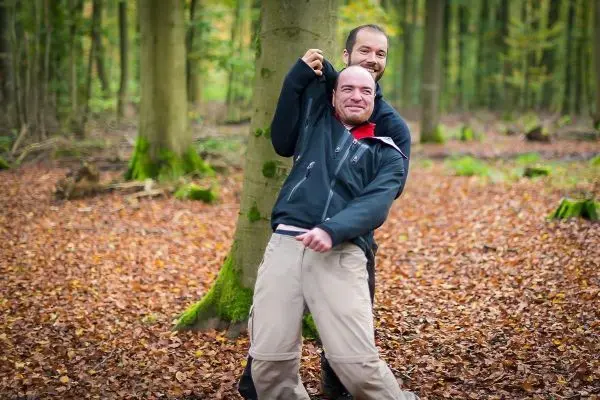 Zwei Personen stehen in einem Wald auf Laub, eine Person hält sich an einem Baum fest