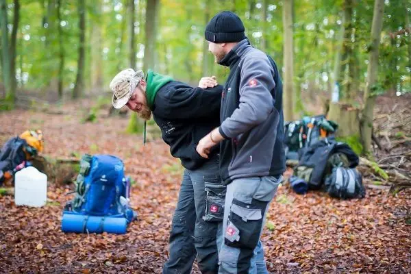 Zwei Personen üben eine Technik zur Unterstützung in einem Waldgebiet mit Rucksäcken im Hintergrund