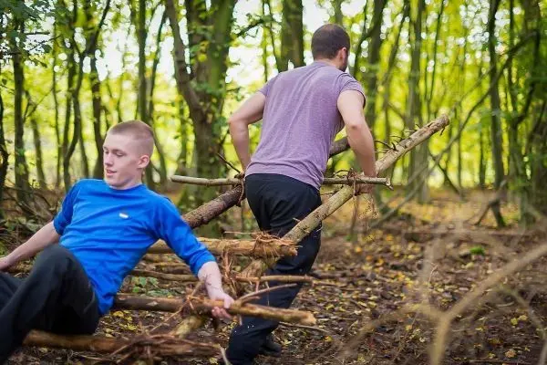Zwei Personen ziehen eine selbstgebaute Konstruktion aus Ästen durch den Wald